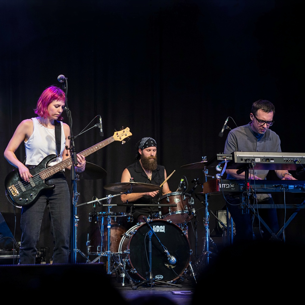 Three musicians on stage: a tall woman with short pink hair 