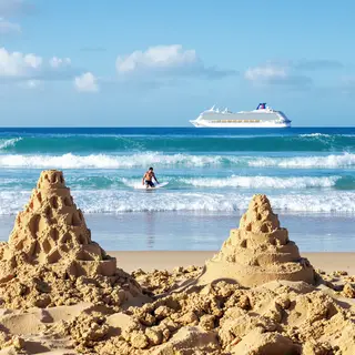 A beach scene with a sandcastle in the foreground, a surfer 