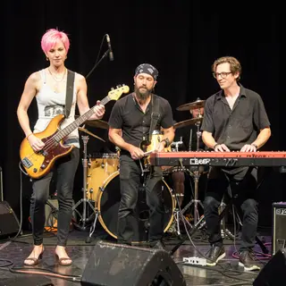 Three musicians on stage: a tall woman with short pink hair 