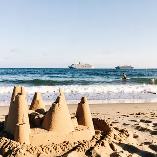 A beach scene with a sandcastle in the foreground, a surfer 