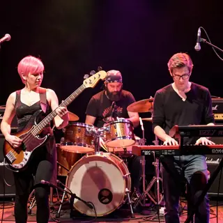 Three musicians on stage: a tall woman with short pink hair 