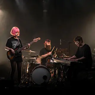 Three musicians on stage: a tall woman with short pink hair 
