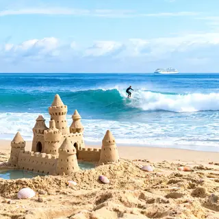 A beach scene with a sandcastle in the foreground, a surfer 