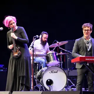Three musicians on stage: a tall woman with short pink hair 