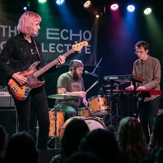 Three musicians on stage: a tall woman with short pink hair 