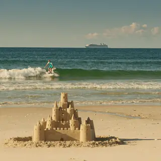 A beach scene with a sandcastle in the foreground, a surfer 