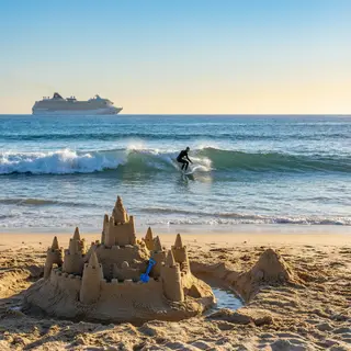 A beach scene with a sandcastle in the foreground, a surfer 