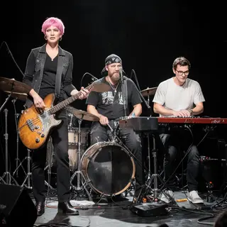 Three musicians on stage: a tall woman with short pink hair 