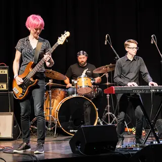 Three musicians on stage: a tall woman with short pink hair 