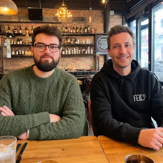 Two men at a cafe: one with a beard and glasses wearing a gr