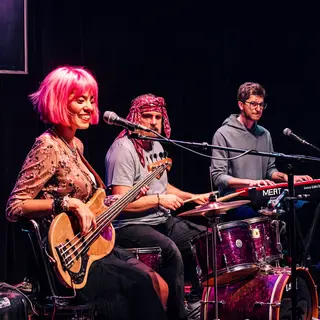 Three musicians on stage: a tall woman with short pink hair 
