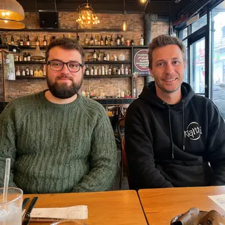 Two men at a cafe: one with a beard and glasses wearing a gr