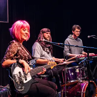 Three musicians on stage: a tall woman with short pink hair 