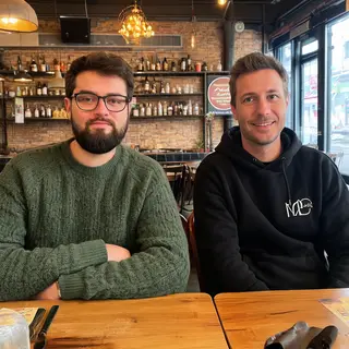 Two men at a cafe: one with a beard and glasses wearing a gr