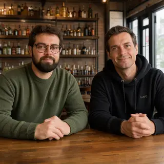 Two men at a cafe: one with a beard and glasses wearing a gr