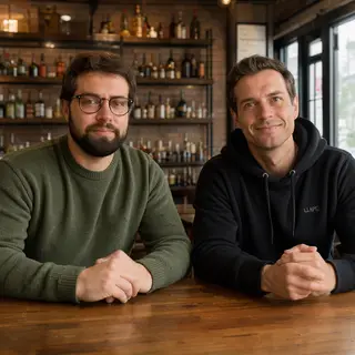 Two men at a cafe: one with a beard and glasses wearing a gr