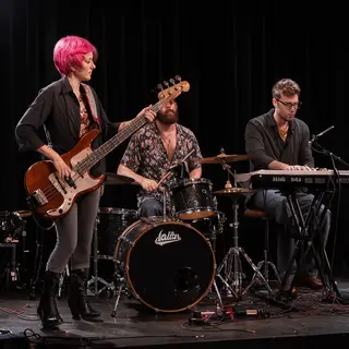 Three musicians on stage: a tall woman with short pink hair 