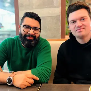 Two men at a cafe: one with a beard and glasses wearing a gr