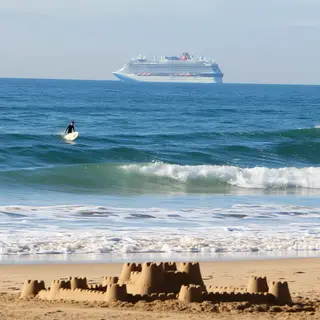A beach scene with a sandcastle in the foreground, a surfer 