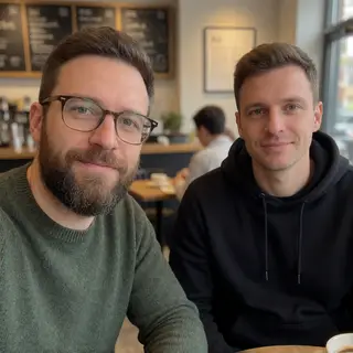 Two men at a cafe: one with a beard and glasses wearing a gr