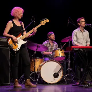 Three musicians on stage: a tall woman with short pink hair 