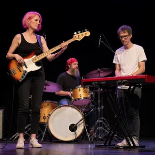 Three musicians on stage: a tall woman with short pink hair 