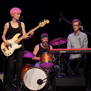 Three musicians on stage: a tall woman with short pink hair 