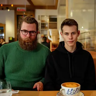 Two men at a cafe: one with a beard and glasses wearing a gr