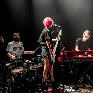 Three musicians on stage: a tall woman with short pink hair 