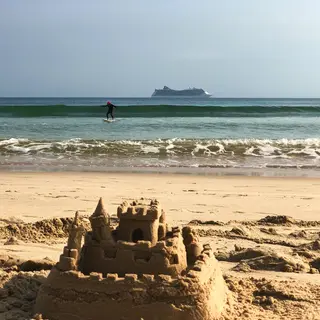 A beach scene with a sandcastle in the foreground, a surfer 