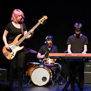 Three musicians on stage: a tall woman with short pink hair 