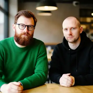 Two men at a cafe: one with a beard and glasses wearing a gr