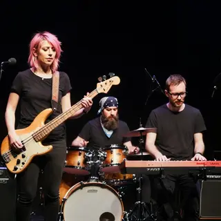 Three musicians on stage: a tall woman with short pink hair 