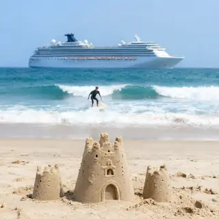 A beach scene with a sandcastle in the foreground, a surfer 