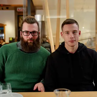 Two men at a cafe: one with a beard and glasses wearing a gr