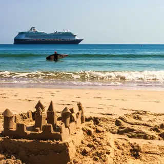 A beach scene with a sandcastle in the foreground, a surfer 