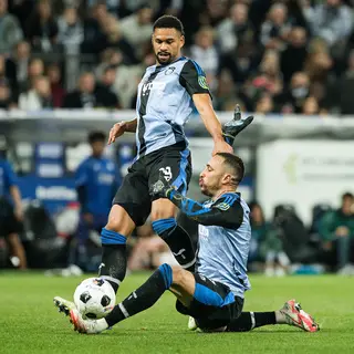 Two soccer players mid-tackle, one sliding and the other jum