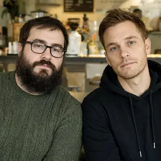 Two men at a cafe: one with a beard and glasses wearing a gr
