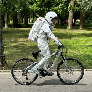 An astronaut in a spacesuit riding a bicycle through a park