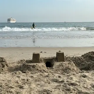 A beach scene with a sandcastle in the foreground, a surfer 