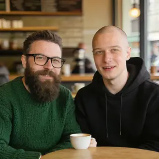 Two men at a cafe: one with a beard and glasses wearing a gr