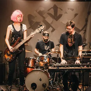 Three musicians on stage: a tall woman with short pink hair 