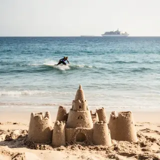 A beach scene with a sandcastle in the foreground, a surfer 