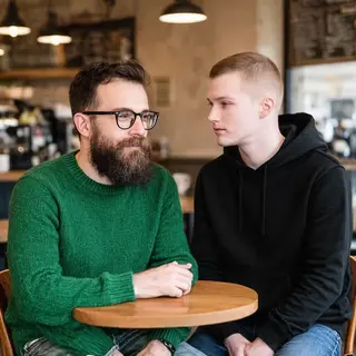 Two men at a cafe: one with a beard and glasses wearing a gr