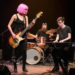Three musicians on stage: a tall woman with short pink hair 