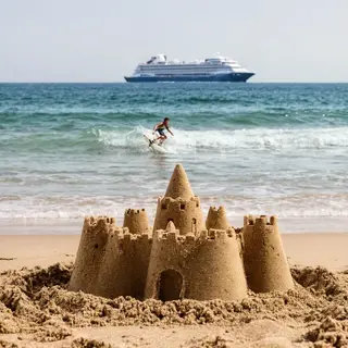 A beach scene with a sandcastle in the foreground, a surfer 