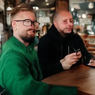 Two men at a cafe: one with a beard and glasses wearing a gr