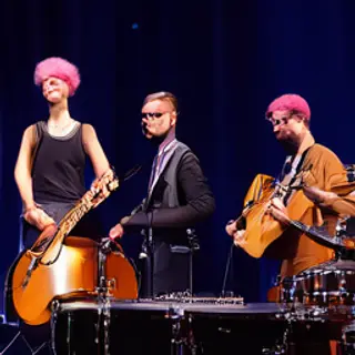 Three musicians on stage: a tall woman with short pink hair 