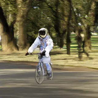 An astronaut in a spacesuit riding a bicycle through a park