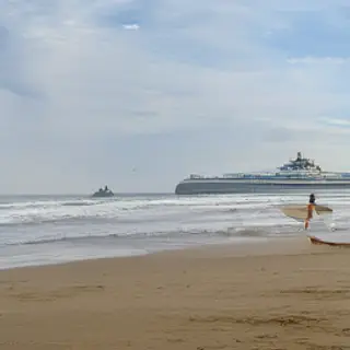 A beach scene with a sandcastle in the foreground, a surfer 